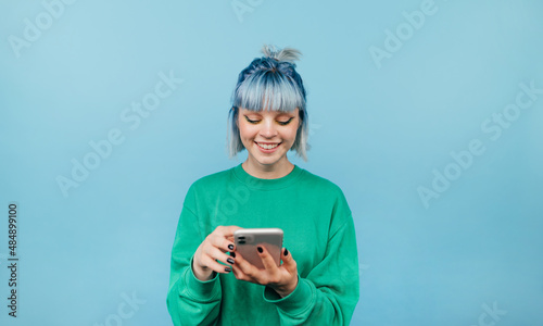 Positive lady with blue hair and green sweatshirt enjoying smartphone on blue background with smile on face looking at screen.