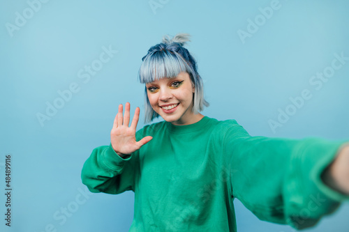 Positive girl with blue hair takes a selfie and smiles at the camera, shows a palm of congratulations, isolated on a blue background.