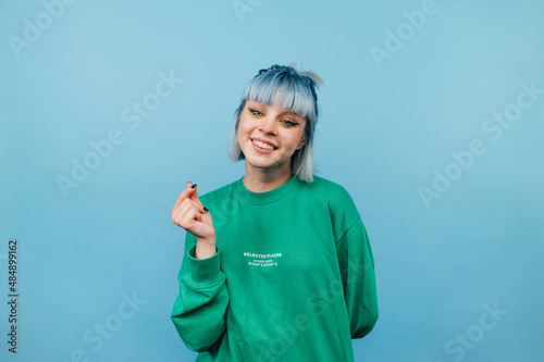 Cute teen girl with colored hair stands on a blue background and with a smile on his face shows his fingers gesture heart