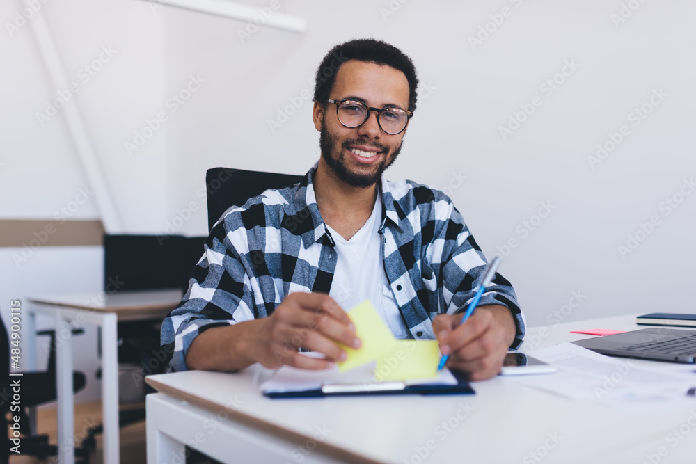 Portrait of happy male executive manager in eyeglasses smiling at camera while creating business plan and strategy for company, student posing at desktop enjoying time for exam Portrait of happy male executive manager in eyeglasses smiling at camera while creating business plan and strategy for company, student posing at desktop enjoying time for exam