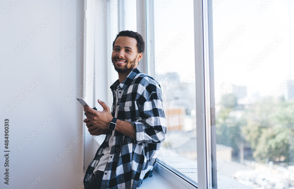 Portrait of happy male blogger in shirt holding mobile phone smiling at camera enjoying daytime indoors, cheerful millennial user with cellphone technology for networking posing near window Portrait of happy male blogger in shirt holding mobile phone smiling at camera enjoying daytime indoors, cheerful millennial user with cellphone technology for networking posing near window