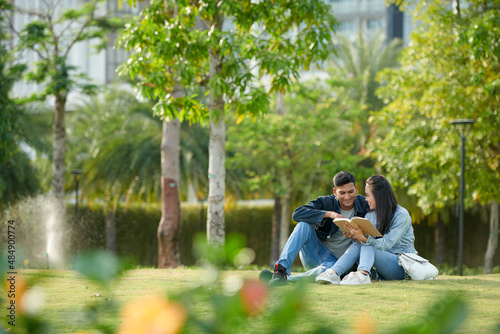 Young university students reading book for class together when sitting on grass on campus