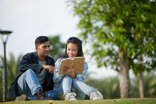Smiling young couple sitting on grass in park and discussing article in non-fictional book