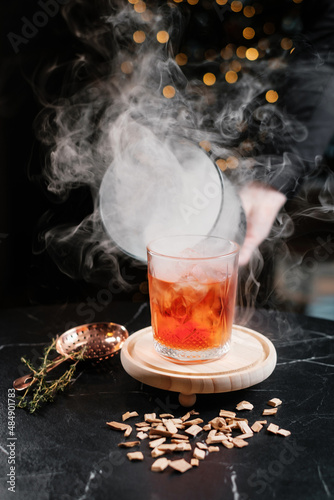 Cocktail served on a wooden board with a glass dome and smoke. The bartender raising the cloche. Concept of aesthetic drinks serving.
