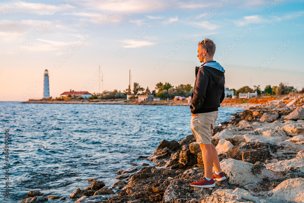 A young blond man looking at the sunset on the sea and the lighthouse, Crimea. Beautiful sea colorful landscape