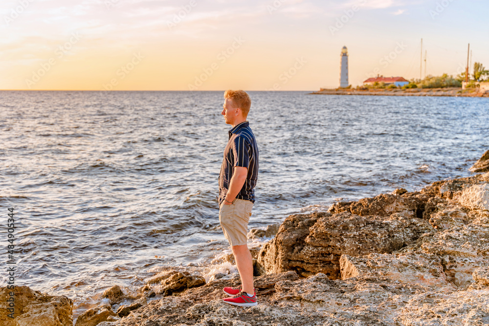 A young blond man looking at the sunset on the sea and the lighthouse, Crimea. Beautiful sea colorful landscape