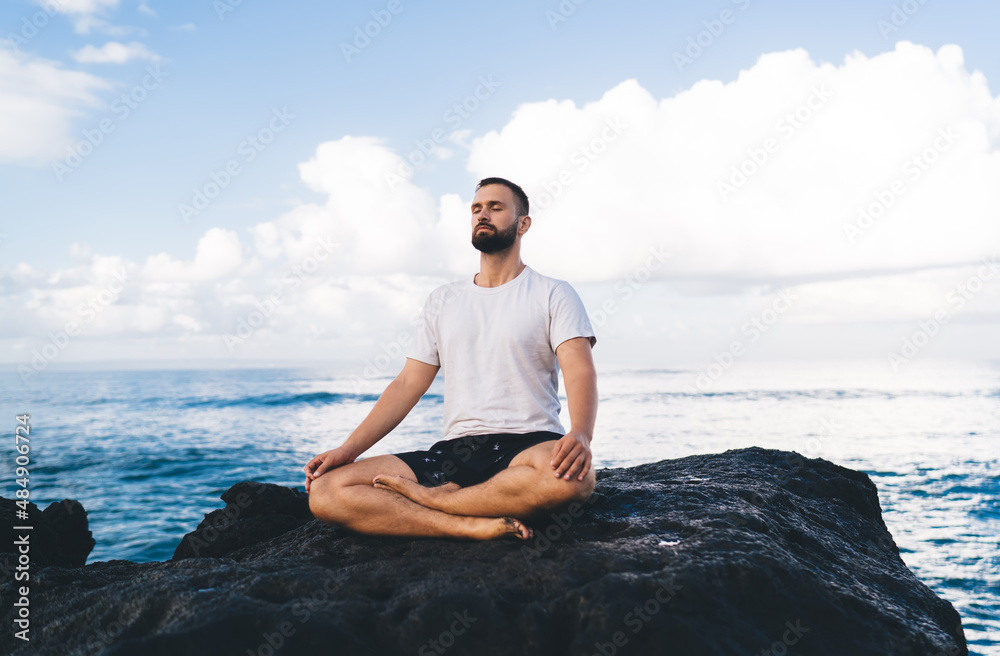 Caucasian male yogi dressed in comfortable wear meditating in lotus pose enjoying relaxation and concentration time at seashore rock, concept of holistic recovery and mental healthy lifestyle Caucasian male yogi dressed in comfortable wear meditating in lotus pose enjoying relaxation and concentration time at seashore rock, concept of holistic recovery and mental healthy lifestyle