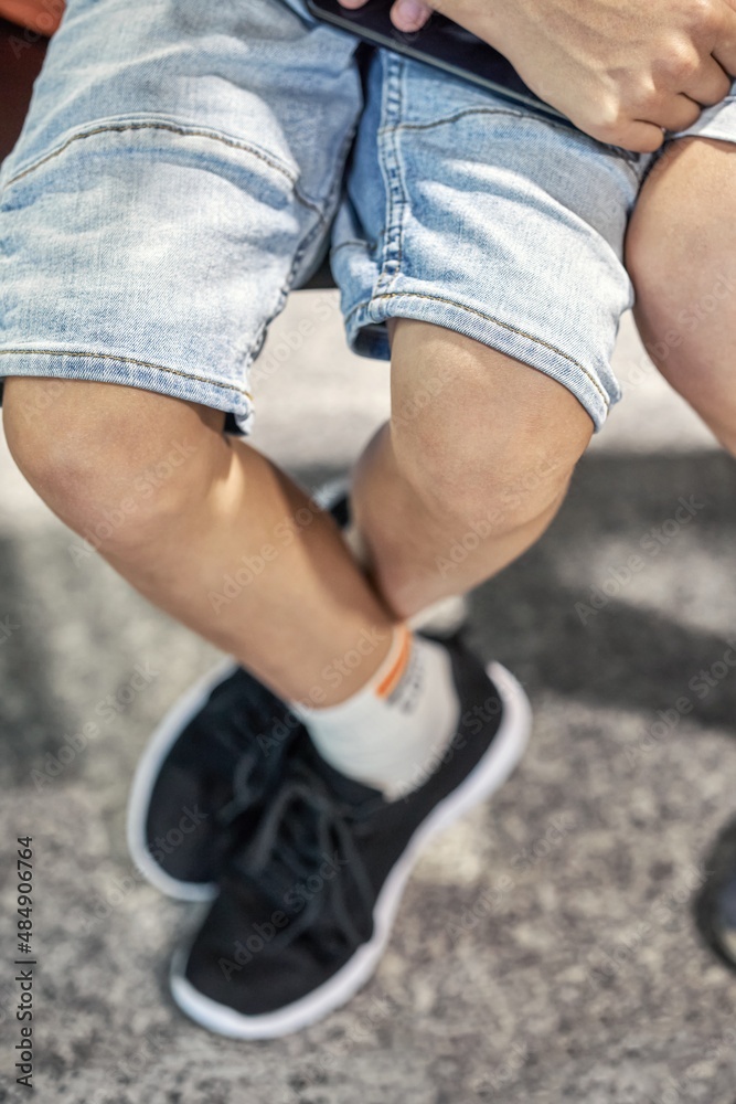 Obraz premium Legs of boy wearing denim shorts sitting on armchair and holding smartphone near brother in waiting room of airport extreme closeup