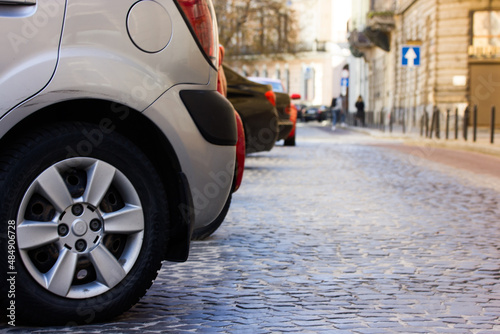 A paved street with cars parked at a curb on a European quiet empty street goes into a distance during a day. Fragment of grey modern car wheel, automobile tire in focus. Parking space. Place for text
