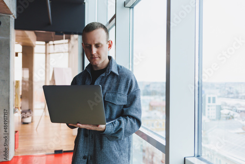 Portrait of a smart intelligent male manager, he is holding a laptop for research in the office, a business man in elegant clothes playing on the touchpad. Standing in the office by a large window © Дмитрий Ткачук