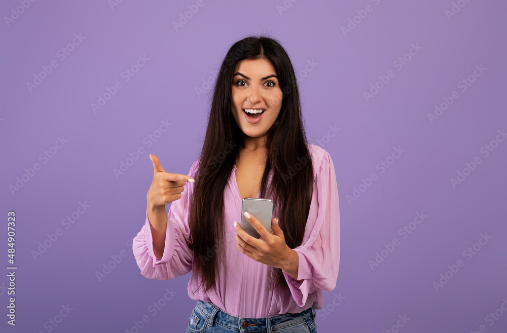 Yes, great news. Surprised armenian woman celebrating win, using smartphone and pointing at gadget, purple background