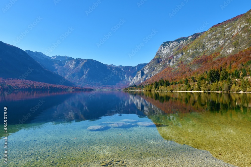 Scenic view of Bohinj lake in Gorenjska, Slovenia with the slopes covered in red colored autumn forest and a reflection of the surrounding mountains in the lake