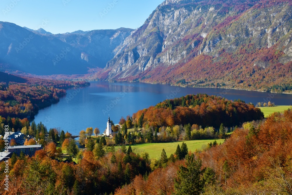 Scenic view of Bohinj lake in Gorenjska, Slovenia in autumn with the forest in red foliage and the mountains of the Julian alps in Triglav national park rising above