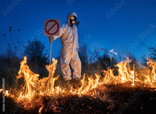 Fireman ecologist extinguishing fire in field at night. Man in protective radiation suit and gas mask near burning grass with smoke, holding prohibition sign. Natural disaster concept.