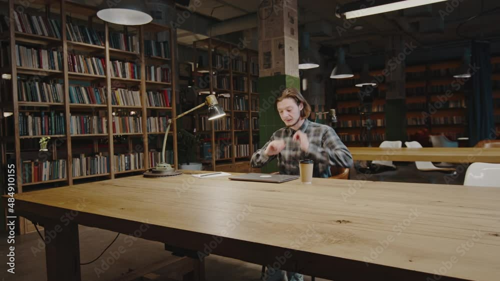 Self education concept. Zoom in shot of young bearded guy studying at library, sitting down at desk and opening laptop Self education concept. Zoom in shot of young bearded guy studying at library, sitting down at desk and opening laptop