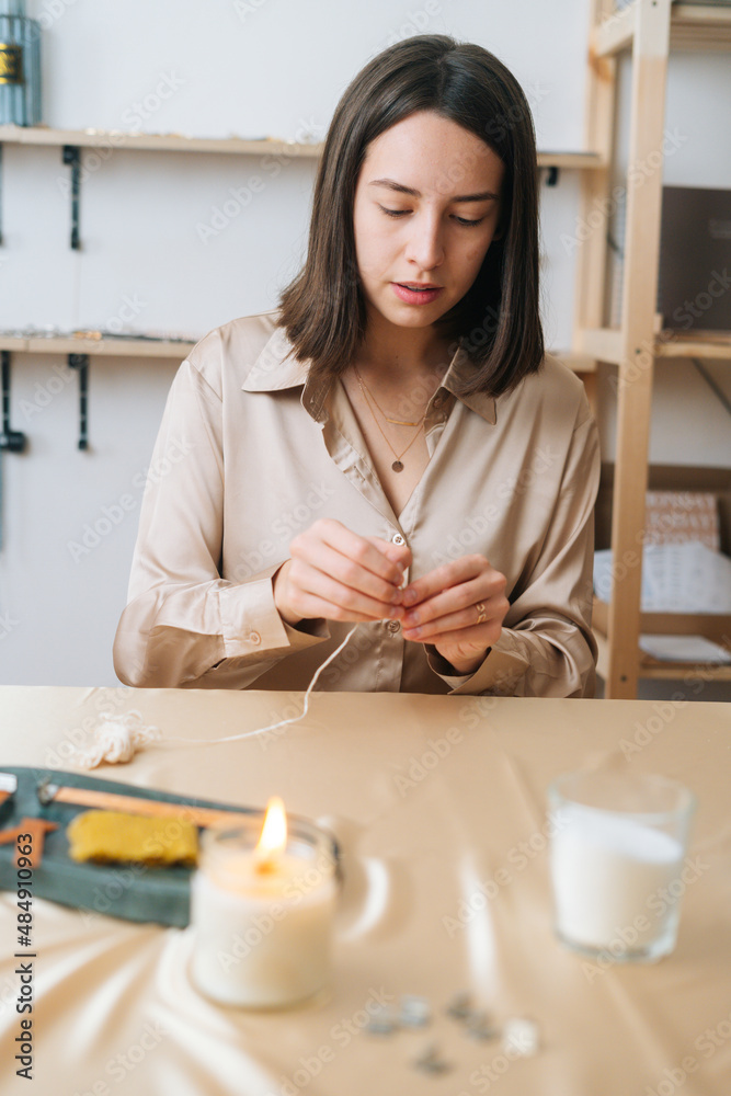 Vertical portrait of focused young woman putting thread through eye of needle at table in modern workshop. Pretty female artisan putting wick through needle. Process of making handmade natural candle Vertical portrait of focused young woman putting thread through eye of needle at table in modern workshop. Pretty female artisan putting wick through needle. Process of making handmade natural candle
