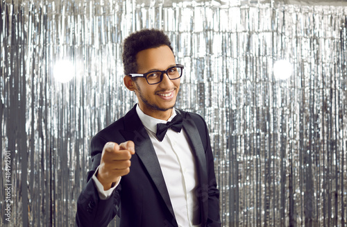 Confident sharp elegant ethnic black man in glasses, chic classic suit, white shirt and bowtie standing on shiny studio background pointing his index finger at camera, smiling and inviting us to party