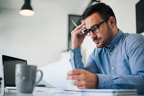 Portrait of stressed out and worried troublesome overworked businessman entrepreneur looking documents report in modern bright office working under pressure and tight deadline photo