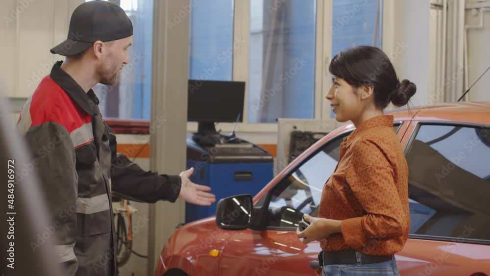 Professional mechanic in uniform giving car key to female client and talking with her in auto service garage