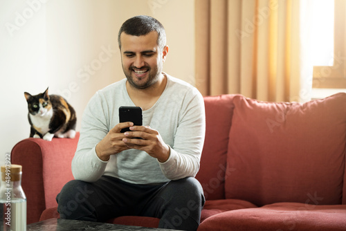 Handsome short haired man holding smartphone and sitting on the couch with his cat. Copy space