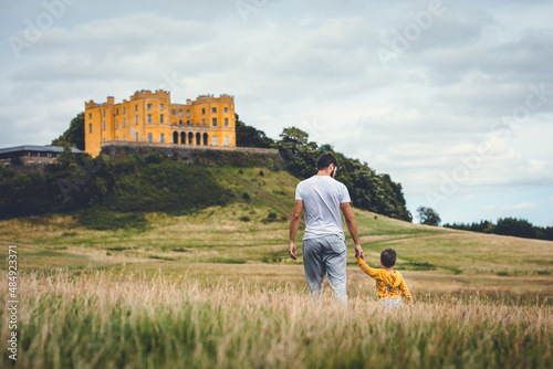 Dad and son walking through a meadow with a castle in the background. Man walking with his son. Yellow castle. Stoke Park Estate, Bristol, UK photo
