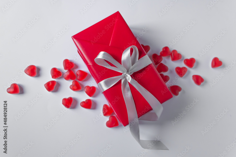 Gift-box is wrapped in red paper with silver ribbon bow. Isolated on a white background in a pile of jelly hearts. Gift-box is wrapped in red paper with silver ribbon bow. Isolated on a white background in a pile of jelly hearts.