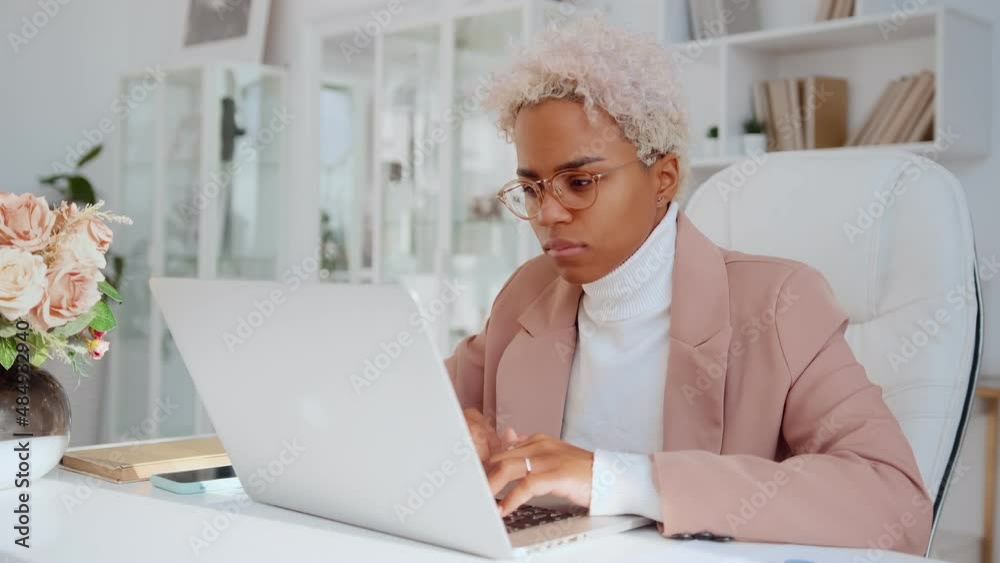 Young successful African American business woman typing text on laptop working on new project staring at computer screen doing reports for own company sits at table in modern office. Young successful African American business woman typing text on laptop working on new project staring at computer screen doing reports for own company sits at table in modern office.