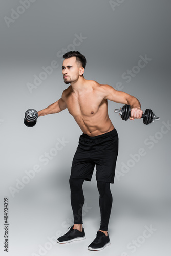 full length of strong sportsman training with dumbbells on grey.