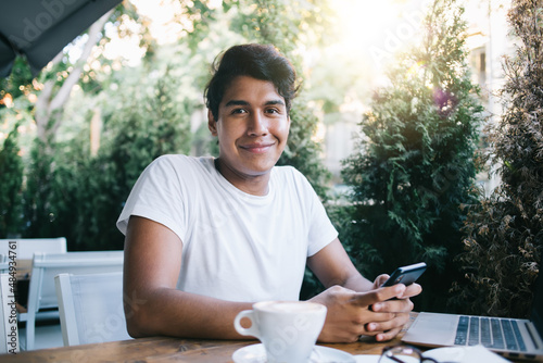 Portrait of millennial blogger holding modern cellphone technology in hands sitting at street cafeteria and smiling at camera, Latino male generation Z enjoying leisure for using devices on freelance