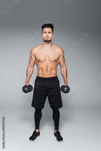 full length of muscular sportsman working out with dumbbells on grey. © LIGHTFIELD STUDIOS