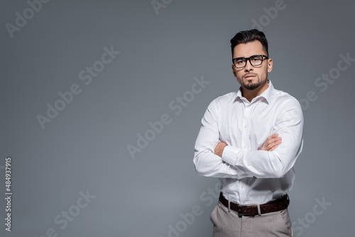 bearded businessman in white shirt and glasses standing with crossed arms isolated on grey. © LIGHTFIELD STUDIOS