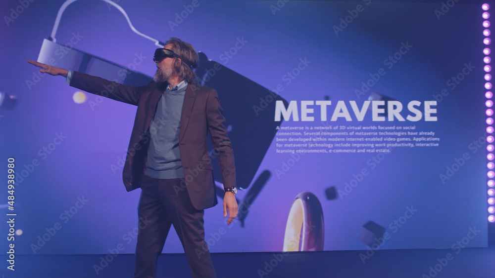 Happy male speaker in suit gesturing and demonstrating augmented reality glasses, to the audience at a Metaverse event on stage in front of an LED screen 3D objects in illuminated room Happy male speaker in suit gesturing and demonstrating augmented reality glasses, to the audience at a Metaverse event on stage in front of an LED screen 3D objects in illuminated room