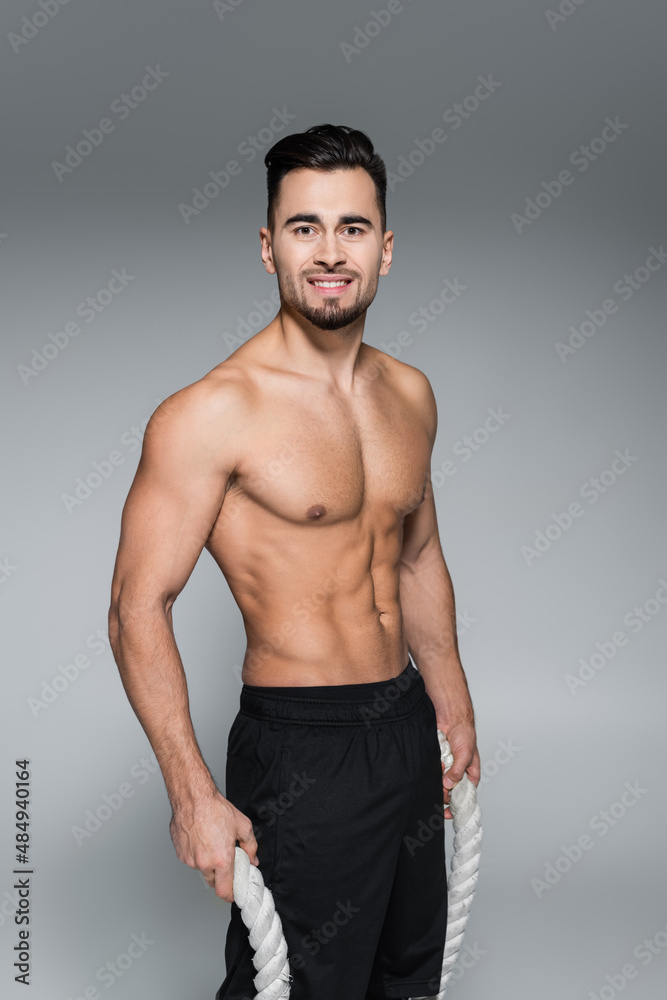 cheerful and shirtless sportsman holding battle ropes on grey.