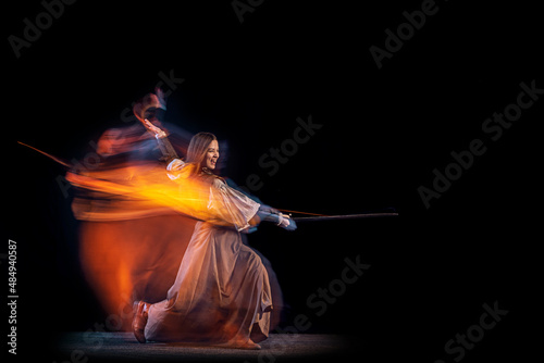 Portrait of beautiful woman in image of medieval warrior or knight with dirty wounded face with big sword isolated over dark background in mixed neon light.