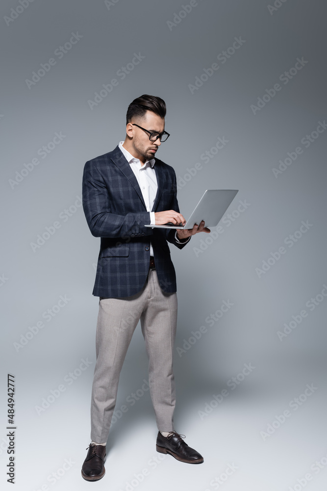 full length of businessman in suit and glasses using laptop on grey.