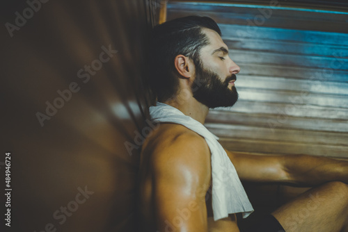 Beautiful healthy boy relaxing in a wooden sauna. Young bearded man resting after workout in the gym. Fitness male with tattoo and white towel, laying on the bench in the spa. Wellness, health concept