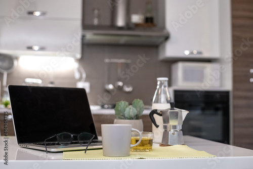 Table set for breakfast, kitchen in the background. Natural meal and technology devices. Interior view of a cozy and modern home. Millennial, youth, home working, home schooling, remote work concept.