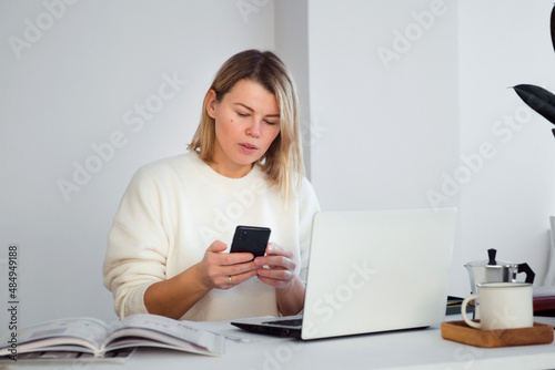 female freelancer online working at table using smartphone, young woman using laptop for business communication or video chatting