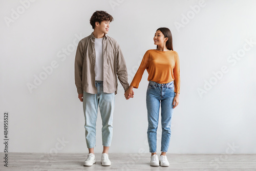 Full length of millennial Asian couple smiling and looking at each other, holding hands, standing against white wall photo