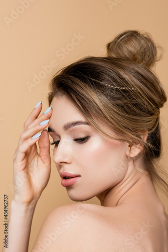 young woman with natural makeup and bare shoulder touching forehead isolated on beige.