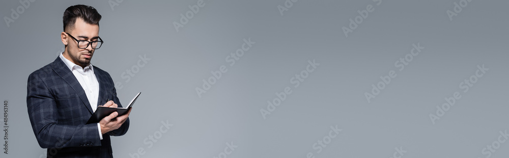 young businessman in suit and glasses writing in notebook isolated on grey, banner. young businessman in suit and glasses writing in notebook isolated on grey, banner.