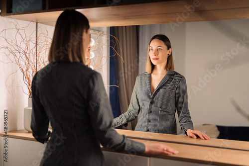 Self-confident Woman looking at her reflection into the mirror indoors. Beautiful interior design