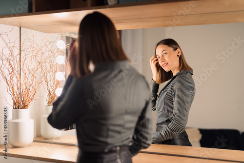 Self-confident Woman looking at her reflection into the mirror indoors. Beautiful interior design