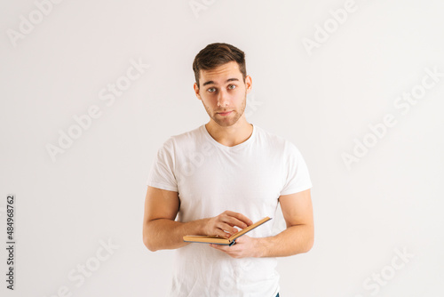 Studio portrait of handsome young man holding in hands paper book on white isolated background in studio, looking at camera. Front view of serious male student studying reading educational materials.