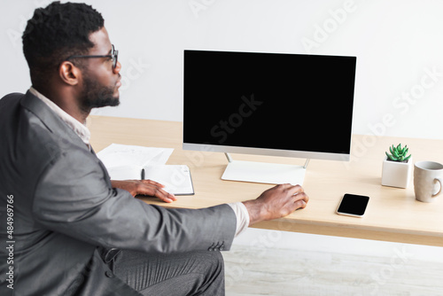 Young black businessman sitting at desk with empty computer monitor, mockup for website design