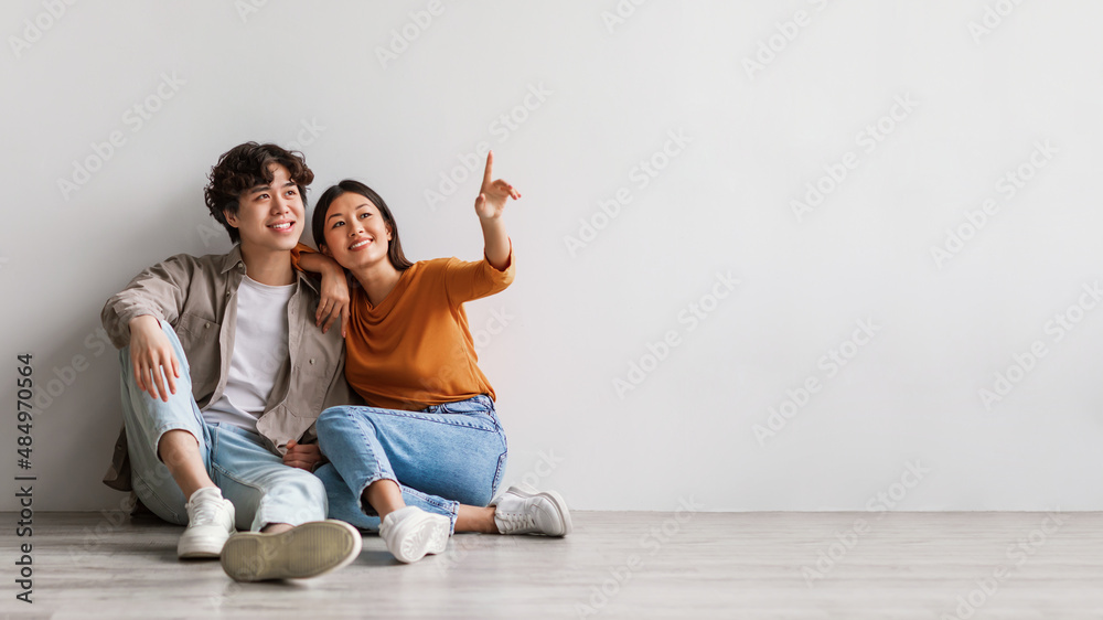 Carefree millennial Asian man and woman sitting on floor near white studio wall and pointing at copy space, banner design Carefree millennial Asian man and woman sitting on floor near white studio wall and pointing at copy space, banner design