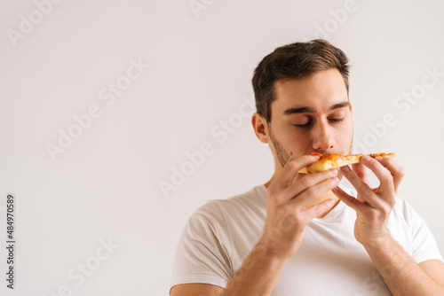 Close-up face of hungry young man enjoying sniffing slice of pizza and eating on delicious food on white isolated background. Studio shot of happy handsome male with closed eyes eating tasty meal.