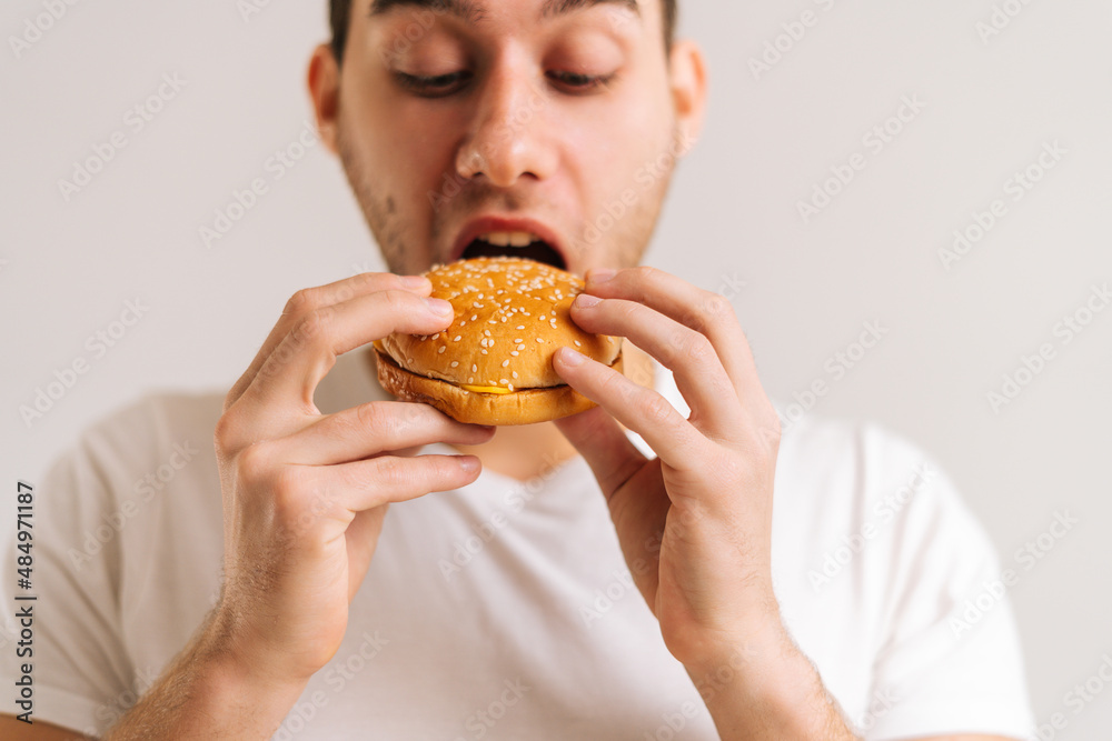 Close-up face of handsome young man bitting eating delicious burger on white isolated background. Studio shot of happy hungry male holding tasty unhealthy meal.