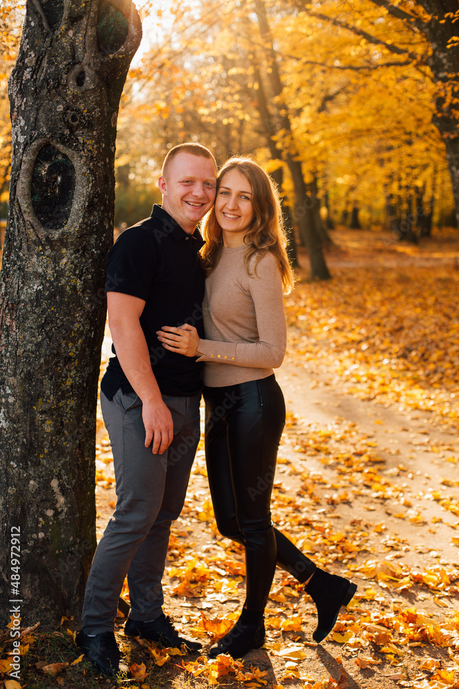 Young couple is having a walk in the sunny autumn park, forest. Happy man and his woman are hugging near yellow tree. Fallen yellow leaves, brith warm autumn weather. Autumn love story in park
