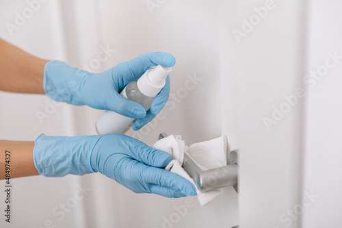 Cleaning black door handle with an antiseptic wet wipe, blue gloves and sanitizer. Sanitize surfaces prevention in hospital and public spaces against corona virus. Woman hand using towel for cleaning.